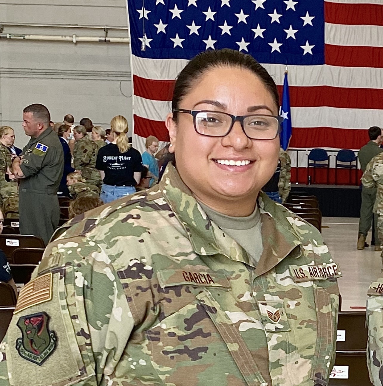 Smiling individual in a U.S. Air Force uniform stands indoors. A large American flag is displayed in the background, with other military personnel nearby.