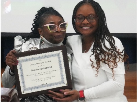 Two individuals pose together, one holding a framed certificate. The certificate reads “Mary Mahoney Award” and honors Kendra Springfield for perseverance in nursing school. Both are smiling in a celebratory setting.