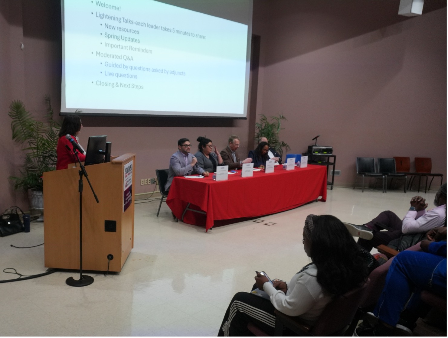 A panel of five speakers at a conference table draped in red, with a woman at a podium beside a large presentation screen displaying agenda topics.