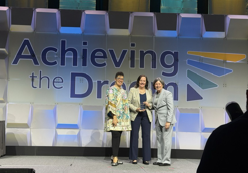 Three individuals stand on a stage in front of a large backdrop that reads “Achieving the Dream.” The backdrop features geometric, three-dimensional wall panels lit with blue and purple lighting. The three individuals stand close together at center stage, appearing to hold an award or document. A table with a dark tablecloth sits to the left, and silhouettes of audience members are visible in the foreground
