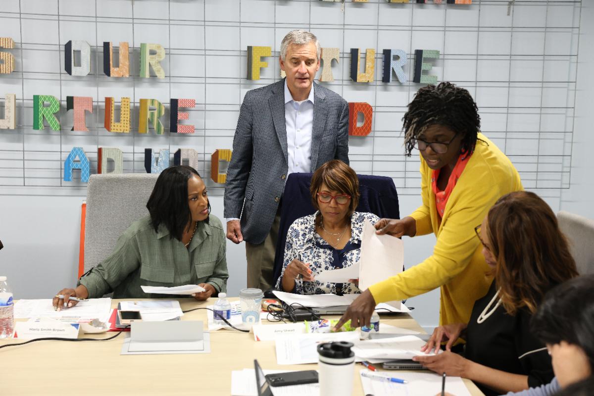 4 individuals sitting at a table looking at papers with one person standing behind them